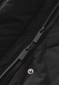 Schwarzer, gesteppter Stoff mit Reißverschluss und metallenen Druckknopfdetails. Verfügt über eine strukturierte Oberfläche und kontrastierende schwarze Hardware-Akzente.