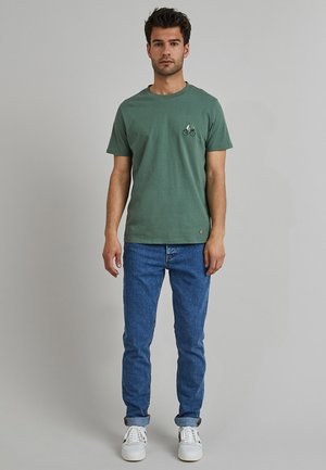 Camiseta de algodón verde con cuello redondo y mangas cortas, que presenta un gráfico de bicicleta en el pecho. Combinada con jeans de mezclilla azules y zapatillas blancas.
