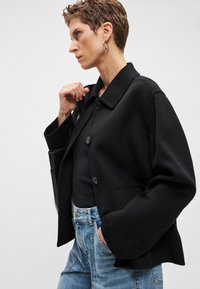 Blusão preto em mistura de lã com gola de colarinho, fecho de botões, ombros caídos e bolsos laterais, combinado com jeans de ganga azul claro.