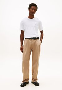 Man staat met handen in de zakken, draagt een wit shirt met korte mouwen, beige geplooide broek, zwarte riem en zwarte schoenen op een witte achtergrond.