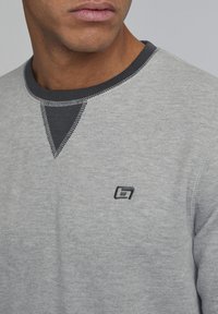 Grauer Sweatshirt mit einem kontrastierenden, dunkelgrauen Rippkragen, ausgestattet mit einem kleinen Logo auf der Brust und sichtbaren Nähdetails.