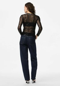 Crna dugmadna bluza s dugim rukavima i transparentnom tkaninom, u kombinaciji s tamnoplavim trapericama visokog struka. Pogled s leđa prikazuje složen uzorak i detalje kroja.