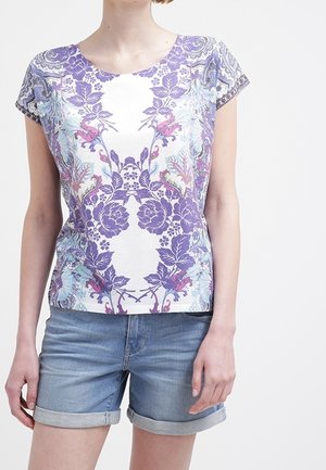 Kobieta w krótkim białym topie z purpurowym wzorem kwiatowym i podwiniętych szortach jeansowych, stojąca na jednolitym białym tle.