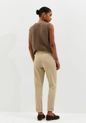 Vrouw met donker haar in een knot, gekleed in een mouwloos bruin bovenstuk, beige opstropende broek en bruine loafers, staand tegen een effen witte achtergrond.