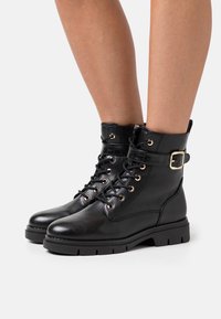 Czarne skórzane botki za kostkę z sznurowanym przodem, złotymi oczkami i bocznym sprzączką. Gruba podeszwa o teksturowanym wykończeniu.