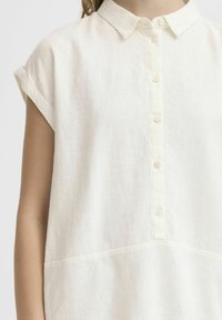 Donna che indossa una camicia button-up a maniche corte di un bianco sporco, con colletto e una cucitura decorativa a metà corpo.