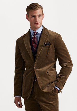 Bruine corduroy pak met een gestructureerde afwerking, knoopsluiting en geknikte revers. Gepaard met een blauwe shirt en een patronen stropdas.
