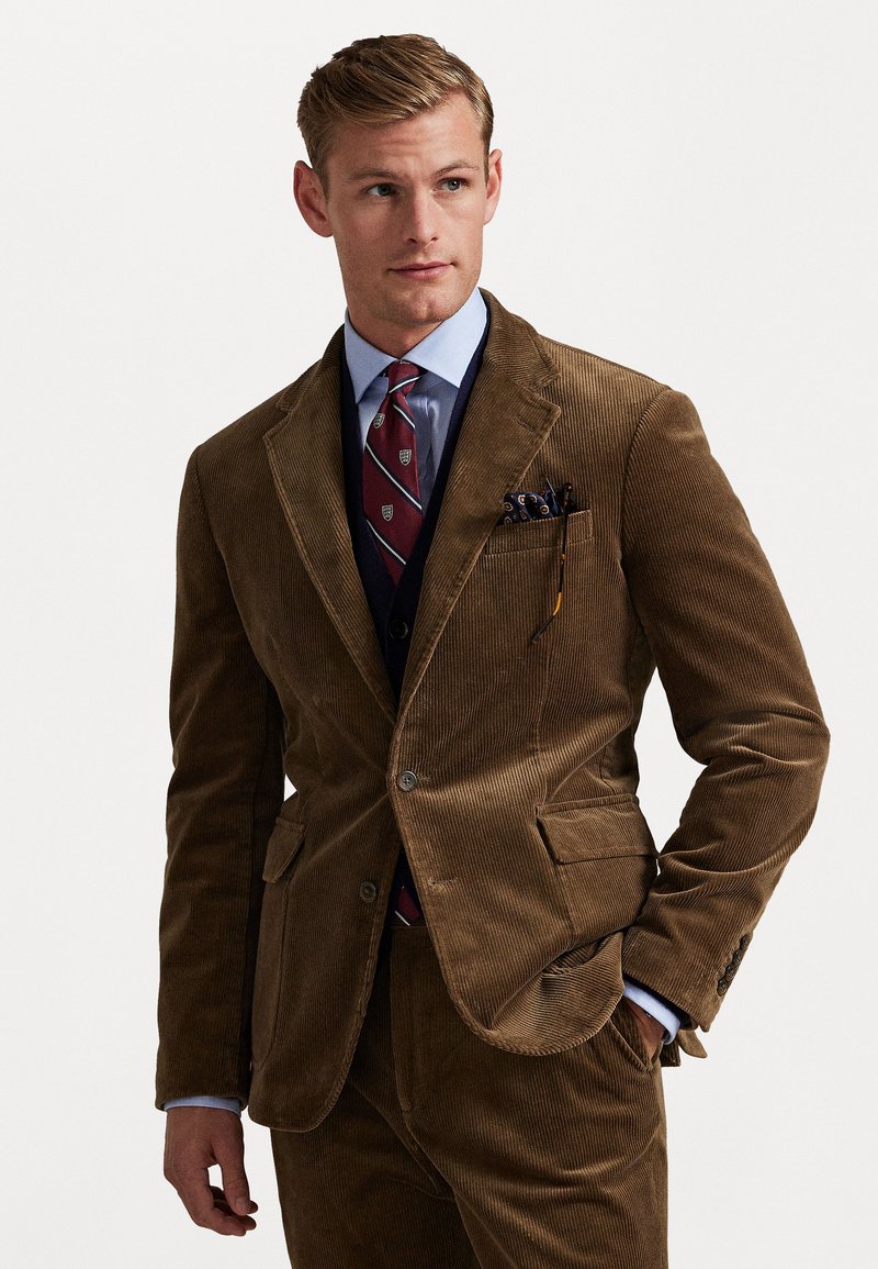 Costume en velours côtelé marron avec une finition texturée, fermeture à boutons et revers classiques. Associé à une chemise bleue et une cravate à motifs.