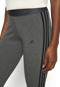 Grå sporttights gjorda av ett slätt, elastiskt material. Har en svart tre-stripesdesign längs sidorna och en märkeslogotyp.