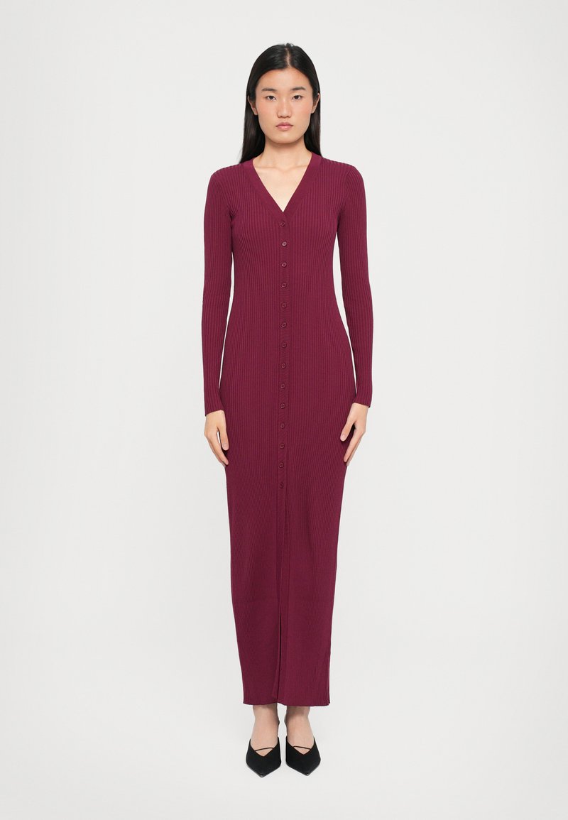 Long burgundy ribbed dress with V-neck, full-length sleeves, vertical button detailing, and a straight silhouette, complemented by black heels.