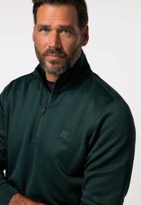 Pullover verde scuro a mezza zip realizzato in materiale liscio e elastico, con un piccolo logo sul petto e dettagli cuciti sulle maniche.