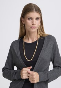 Pilkas kardiganas su atvira priekiu, dryžuotais kraštais, dėvimas ant juodo viršaus. Papuoštas auksine karoliukų vėriniu. Lygus tekstūros, ilgomis rankovėmis. Kasdienio stiliaus.