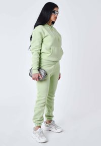 Sudadera y pantalones de chándal a juego en verde claro, de suave material de felpa, con un bolsillo frontal y puños elásticos. Bolsa cruzada plateada y zapatillas blancas.