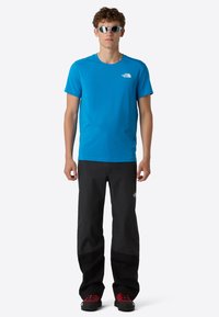 Modrá košile s krátkým rukávem a malým bílým logem, kombinovaná s černými prodyšnými kalhotami a odolnými červenými a černými botami, vhodná pro venkovní aktivity.