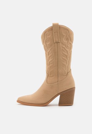 Tan western-style ankle boot with a pointed toe, decorative stitching, and a textured stacked heel. Suede material with a rear pull tab.