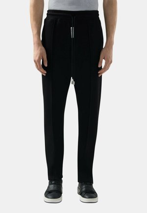 Uomo che indossa pantaloni neri con coulisse e fondo stretto e sneaker nere con suola bianca, in piedi contro uno sfondo chiaro uniforme.