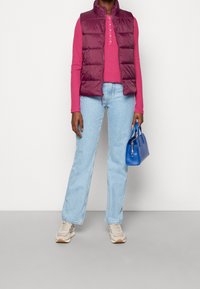 Maroon vadderad väst över en rosa långärmad topp, ljusblå utsvängda jeans, grå sneakers och en liten blå handväska. Släta ytor, avslappnad design.