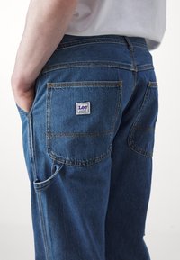 Personne portant un jean bleu Lee Workwear avec une poche arrière visible et un détail de boucle latérale, main reposant dans la poche avant.