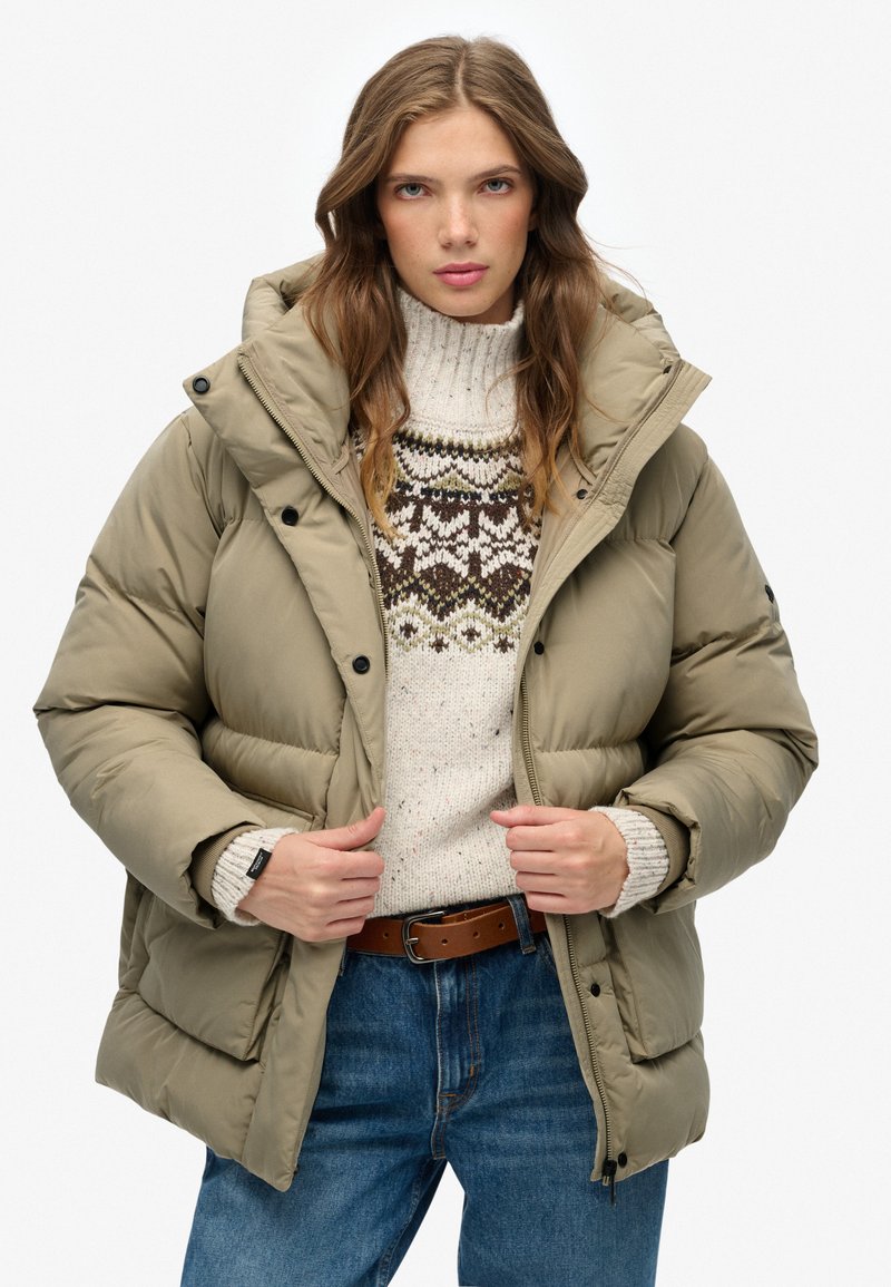 Chaqueta de color caqui acolchada con un cuello alto y botones negros, superpuesta a un suéter crema con estampado. Unos vaqueros azules y un cinturón marrón completan el look.