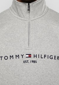 Sweat-shirt gris clair à fermeture éclair quart avec le logo « Tommy Hilfiger » et un emblème de drapeau rouge, blanc et bleu brodé au-dessus du texte « EST. 1985 ».