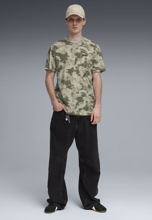 Junger Mann mit beiger Puma-Kappe, Puma-T-Shirt mit Tarnmuster, lockeren schwarzen Jeans mit Schlüsselanhänger und schwarzen Sneakers vor grauem Hintergrund.