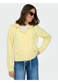 Femme souriante portant une veste à capuche jaune avec cordons, des lunettes de soleil, un t-shirt blanc et un jean bleu, posant avec la main sur la hanche.
