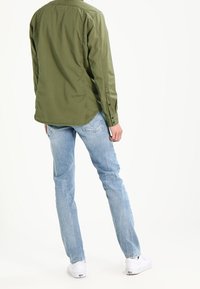 Camicia a maniche lunghe verde oliva, abbottonata, abbinata a pantaloni in denim azzurro chiaro. Le scarpe sono bianche, con una texture liscia e un design semplice.