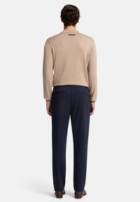Beiger Strickpullover mit langen Ärmeln, kombiniert mit marineblauen Hosen. Die Hose verfügt über zwei Gesäßtaschen, und der Pullover hat ein Logodetail auf der Rückseite.