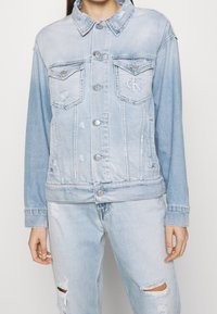 Giacca di jeans azzurro chiaro con colletto classico, due tasche sul petto, chiusura con bottoni e dettagli strappati su maniche e orlo. Logo CK sulla tasca.