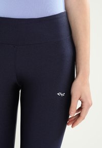 Marinblå leggings med en slät yta, hög midja och en liten vit logga på nedre högra sidan. Tillverkade av ett stretchmaterial.