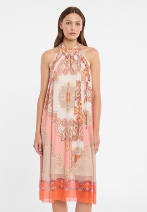 Frau trägt ärmelloses Midikleid mit Neckholder und Paisley- sowie Blumenmustern in Pink, Orange und Beige, steht vor weißem Hintergrund.