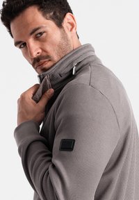 Giacca grigia con zip, superficie testurizzata, colletto alto e una patch nera con logo sulla manica. Presenta dettagli in cerniera argento.