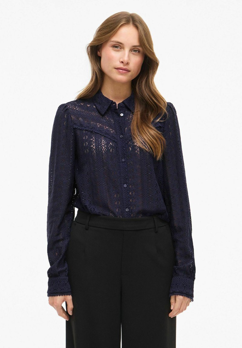 Marineblå blondebluse med knaplukning foran, krave og lange ærmer, der har udsøgte mønstre og en semi-gennemsigtig tekstur.