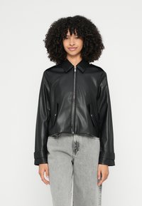 Īsa melna mākslīgās ādas jaka ar priekšējo rāvējslēdzēju, lielu apkakli un sānu kabatām. Kombinēta ar gaiši pelēkām džinsa biksēm.