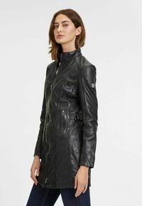 Giacca in pelle nera con colletto alto, chiusura con zip e tasche laterali. Caratterizzata da una vita con cintura e una texture liscia e lucida.