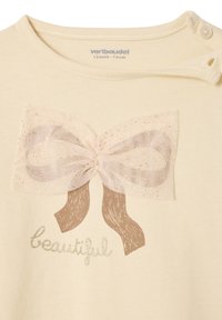 Eine cremefarbene Bluse mit einer Netzschleife in hellrosa und braunen Bändern. "Beautiful" ist in glitzerndem Gold unterhalb der Schleife aufgedruckt.
