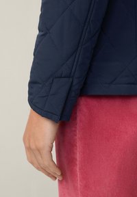 Manica di una giacca in nylon trapuntato blu navy con un polsino leggermente girato, che rivela una texture liscia; mano appoggiata vicino a un indumento in corduroy rosa.