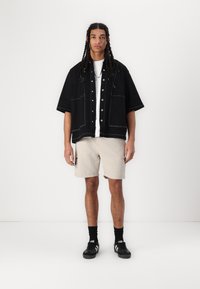 Svart short-sleeve jakke med kontrastfarget hvit søm, hvit T-skjorte, beige cargo-shorts og svarte sneakers med hvite striper.