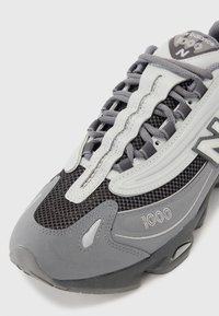 Gros plan sur une chaussure de sport New Balance 1000 grise et noire, montrant la tige en maille, les coutures détaillées, les lacets et le logo sur le côté et la languette.