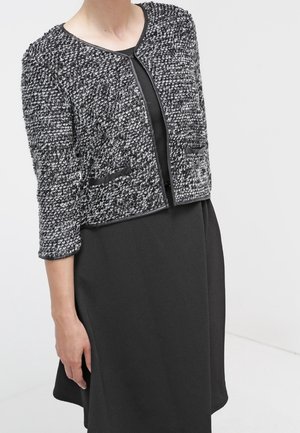 Vrouw draagt een gestructureerde zwart-witte cropped cardigan over een mouwloze zwarte jurk tegen een effen lichte achtergrond.