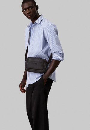 Black crossbody bag made of smooth material with a zippered compartment, worn over a blue and white striped shirt, paired with black trousers.