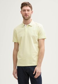 Polo-shirt jaune clair à manches courtes, avec patte de boutonnage à deux boutons et petit logo sur la poitrine gauche. Associé à un jean bleu foncé.