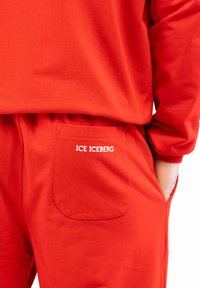 Raudonas sportinis kostiumas, kuriame yra ilgomis rankovėmis marškiniai ir laisvi kelnės. Kelnės turi kišenę su balta siuvinėta užrašu "ICE ICEBERG."