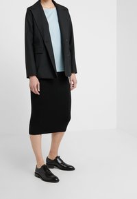 Blazer noir à revers crantés, haut bleu clair, jupe noire tricotée et chaussures noires à lacets. Texture lisse et coupe ajustée.