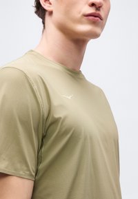 Νέος άνδρας που φοράει μια μπεζ αθλητική μπλούζα με κοντά μανίκια και υφή, με μικρό λευκό λογότυπο στο στήθος, κομμένη στον λαιμό και στους ώμους.