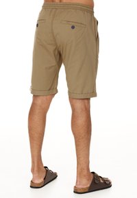 Khaki-Shorts mit lockerem Schnitt, elastischem Bund, umgeschlagenen Säumen und zwei Gesäßtaschen, die mit dunkelblauen Knöpfen gesichert sind. Braune Slipper.