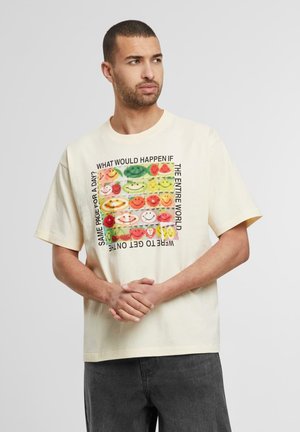 Cremefarvet bomulds T-shirt med en afslappet pasform, prydet med et farverigt frugt- og smiley-ansigt grafik på forsiden. Tekst omgiver designet.