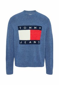 Blå stickad tröja med en stor rektangulär logotyp som har texten "TOMMY JEANS" i vitt, rött och marinblått. Rund halsringning.