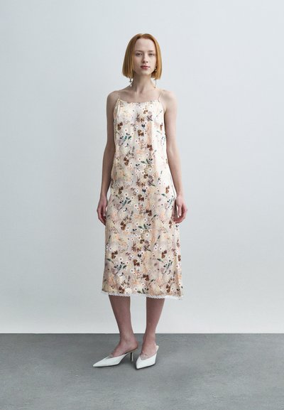 Robe nuisette fleurie en tissu doux, présentant un fond pâle avec une végétation colorée et des bordures en dentelle. Complétée par des mules blanches à bout pointu.