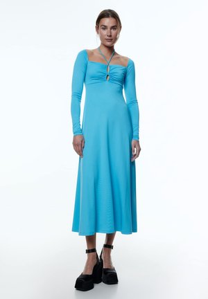 Blaues langärmeliges Kleid mit quadratischem Ausschnitt, zentralem Raffung und einem Schlitzdetail. Fließender Stoff reicht bis zur Mitte der Wade. Schwarze Plateauschuhe.